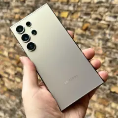 En hand håller en ljusgrå Samsung S24 med flera kameror mot en tegelvägg.