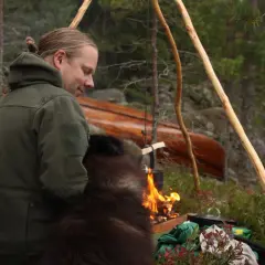 En man med en hund sitter vid en lägereld i skogen. En kanot ligger i bakgrunden. Detta är vad en fotosession betyder för dem.