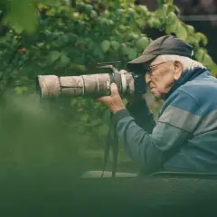Äldre man med keps och glasögon fotograferar med en stor kamera i en lummig trädgård. En passionerad naturfotograf i sitt esse.