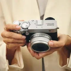 Händerna håller en silverfärgad Fujifilm GFX 100s kamera med ett objektiv.