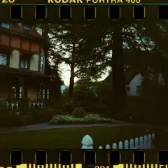 Ett gammalt hus med veranda, omgivet av träd och en trädgård. Bilden har en tydlig film format-känsla med perforeringar.