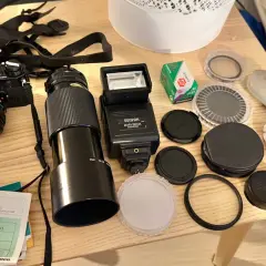 En Canon AE-1 kamera med objektiv, blixt, film och tillbehör ligger på ett träbord.