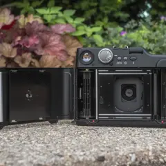 En öppen Fuji GA645 med filmfack och lins synliga. Kameran ligger på en stenbelagd yta med grönska i bakgrunden.