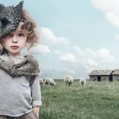 Ett barn med en vargmask i handen, klädd i fuskpäls, står på en äng med får. En saga om Wendotte.