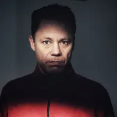 Knut Koivisto, en man med intensiv blick, bär en röd jacka. Ljuset faller dramatiskt över hans ansikte.