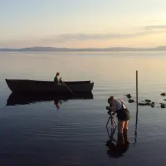 Nygårds Karin Bengtsson filmar en person i en roddbåt vid solnedgången på en lugn sjö.