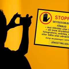 Silhuett av person som fotograferar. Gul skylt med texten "STOPP SKYDDSOBJEKT FÖRBUD" och symboler som varnar för att fotografera, avbilda eller mäta utan tillstånd.
