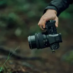 En hand håller en regnvåt Fujifilm GFX 50S II med ett zoomobjektiv i skogen.