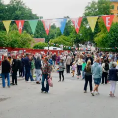 Färgglada vimplar med texten "Planket Stockholm" hänger över en livlig utomhusutställning med många besökare.