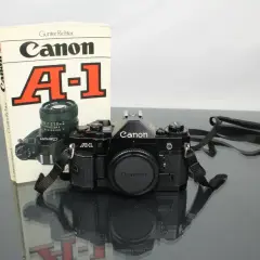 Svart Canon A-1 systemkamera med objektiv och en bok om kameran. En klassiker för den som vill lära sig mer om analog fotografering.