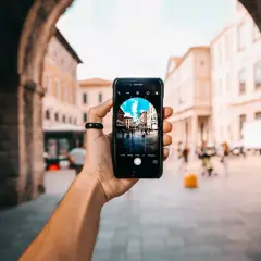 En hand håller upp en iPhone för att ta bra bilder med iPhone av en livlig stadsgata sedd genom en stenbåge.