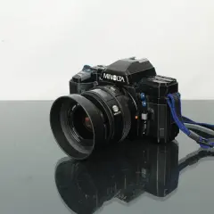 Svart Minolta 7000 systemkamera med objektiv och blå axelrem, placerad på en reflekterande yta.