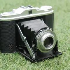 En klassisk Agfa Isolette hopfällbar kamera med läderdetaljer och en silverfärgad lins, placerad på grönt gräs.