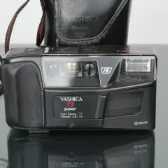 Svart kompaktkamera, en Yashica T3 Super med Carl Zeiss Tessar-objektiv, med en svart läderfodral och en röd-svart kamerarem.