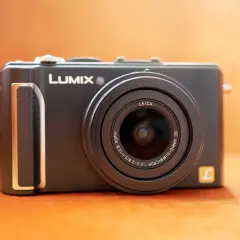 Svart kompaktkamera, en Lumix LX3 med Leica-objektiv, ligger på en orange yta.