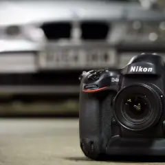 En Nikon D4S-kamera med objektiv står på ett bord. I bakgrunden skymtar en bil.