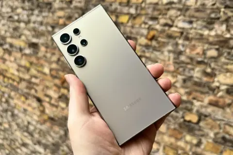 En hand håller en ljusgrå Samsung S24 med flera kameror mot en tegelvägg.