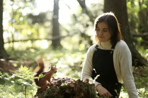 En ung kvinna, Dani Connor, sitter i skogen och tittar på en ekorre som sitter på en stubbe.