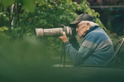 Äldre man med keps och glasögon fotograferar med en stor kamera i en lummig trädgård. En passionerad naturfotograf i sitt esse.