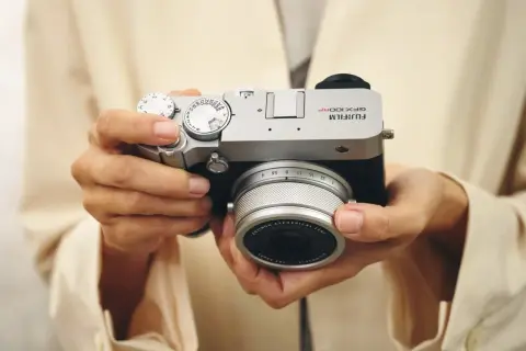 Händerna håller en silverfärgad Fujifilm GFX 100s kamera med ett objektiv.