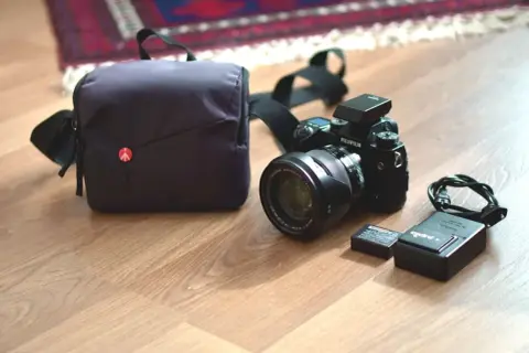 En Fujifilm XH1-kamera med objektiv, batteri, laddare och väska ligger på ett trägolv.