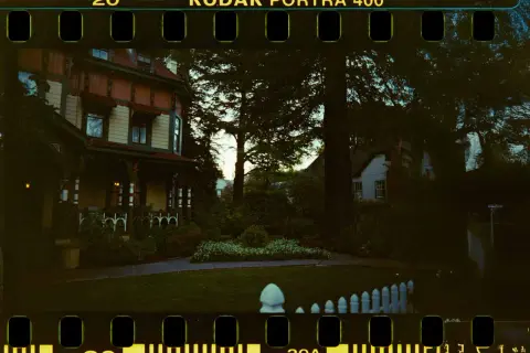 Ett gammalt hus med veranda, omgivet av träd och en trädgård. Bilden har en tydlig film format-känsla med perforeringar.
