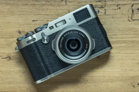 En Fujifilm X100T-kamera ligger på en träyta. Dess retrodesign och fasta objektiv gör den till en favorit för gatufotografering.
