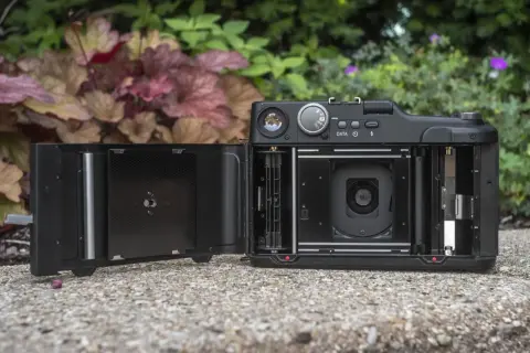 En öppen Fuji GA645 med filmfack och lins synliga. Kameran ligger på en stenbelagd yta med grönska i bakgrunden.