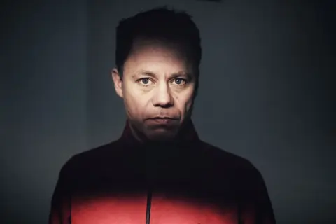 Knut Koivisto, en man med intensiv blick, bär en röd jacka. Ljuset faller dramatiskt över hans ansikte.