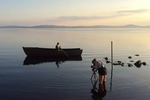 Nygårds Karin Bengtsson filmar en person i en roddbåt vid solnedgången på en lugn sjö.