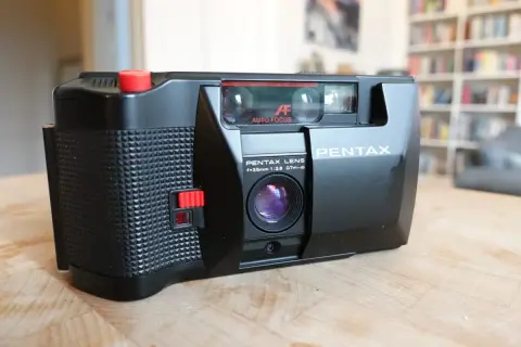 En svart Pentax PC35AF-kamera med autofokus och en 35mm lins ligger på ett träbord.