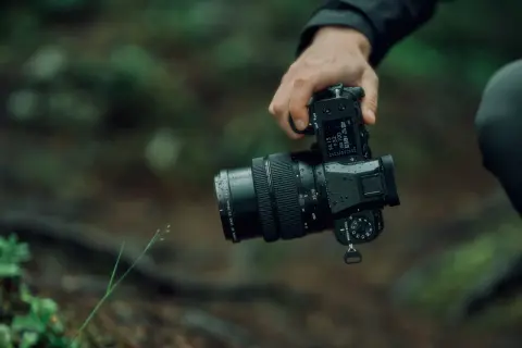 En hand håller en regnvåt Fujifilm GFX 50S II med ett zoomobjektiv i skogen.