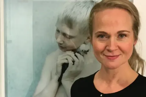 Anna Claren ler mot kameran, med en konstnärlig bild av en pojke som håller en fågel i bakgrunden.