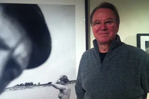 Man i glasögon ler vid en utställning med anders zorn fotografier. En närbild av ett ansikte och en kvinna vid vatten syns i bakgrunden.