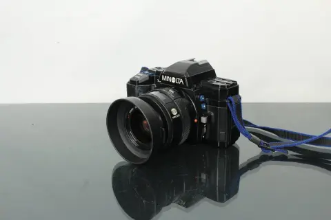 Svart Minolta 7000 systemkamera med objektiv och blå axelrem, placerad på en reflekterande yta.