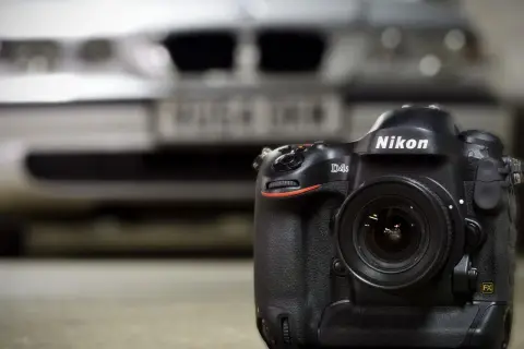 En Nikon D4S-kamera med objektiv står på ett bord. I bakgrunden skymtar en bil.