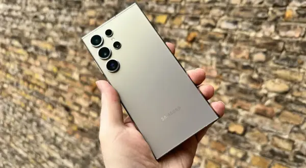 En hand håller en ljusgrå Samsung S24 med flera kameror mot en tegelvägg.