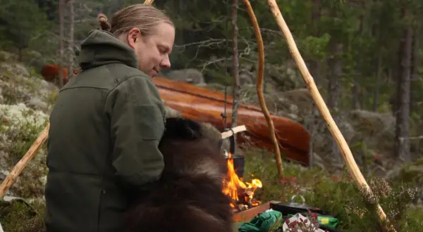 En man med en hund sitter vid en lägereld i skogen. En kanot ligger i bakgrunden. Detta är vad en fotosession betyder för dem.