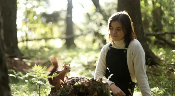 En ung kvinna, Dani Connor, sitter i skogen och tittar på en ekorre som sitter på en stubbe.