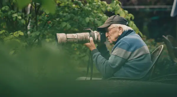 Äldre man med keps och glasögon fotograferar med en stor kamera i en lummig trädgård. En passionerad naturfotograf i sitt esse.