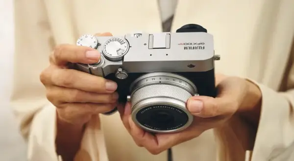 Händerna håller en silverfärgad Fujifilm GFX 100s kamera med ett objektiv.