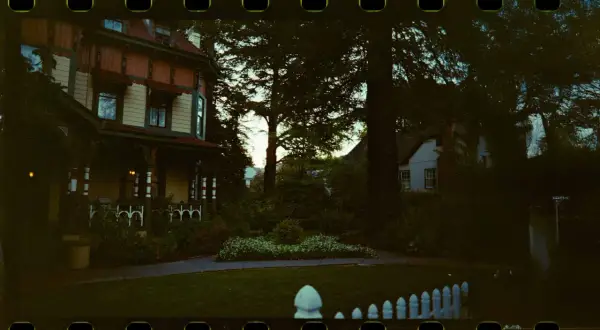 Ett gammalt hus med veranda, omgivet av träd och en trädgård. Bilden har en tydlig film format-känsla med perforeringar.