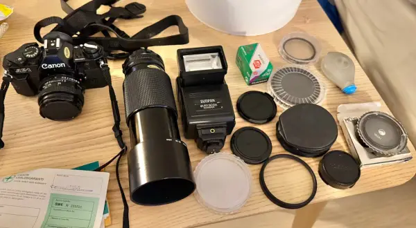 En Canon AE-1 kamera med objektiv, blixt, film och tillbehör ligger på ett träbord.