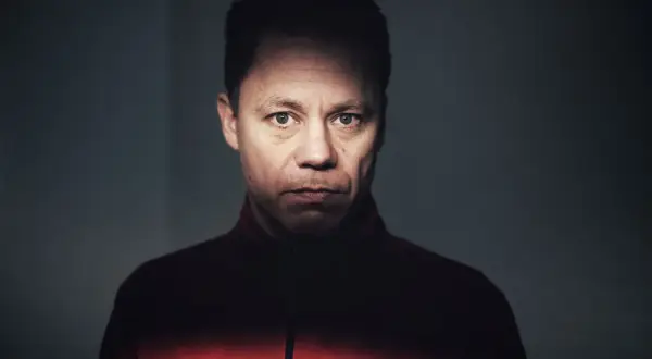 Knut Koivisto, en man med intensiv blick, bär en röd jacka. Ljuset faller dramatiskt över hans ansikte.