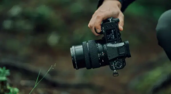 En hand håller en regnvåt Fujifilm GFX 50S II med ett zoomobjektiv i skogen.