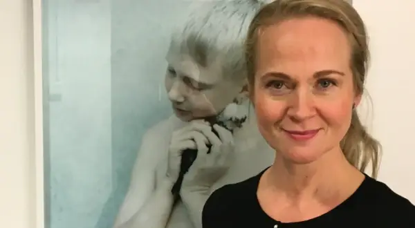 Anna Claren ler mot kameran, med en konstnärlig bild av en pojke som håller en fågel i bakgrunden.