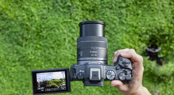En hand håller en Canon systemkamera med objektivet 24-50mm. Kamerans skärm visar en grönskande vy.
