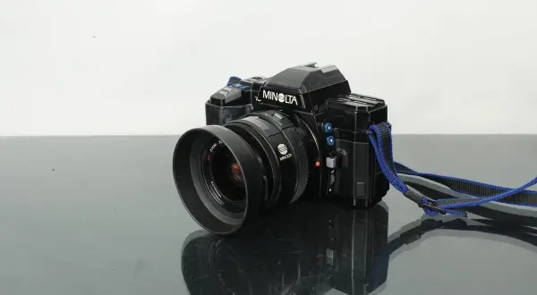 Svart Minolta 7000 systemkamera med objektiv och blå axelrem, placerad på en reflekterande yta.