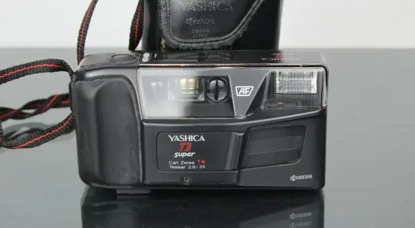 Svart kompaktkamera, en Yashica T3 Super med Carl Zeiss Tessar-objektiv, med en svart läderfodral och en röd-svart kamerarem.