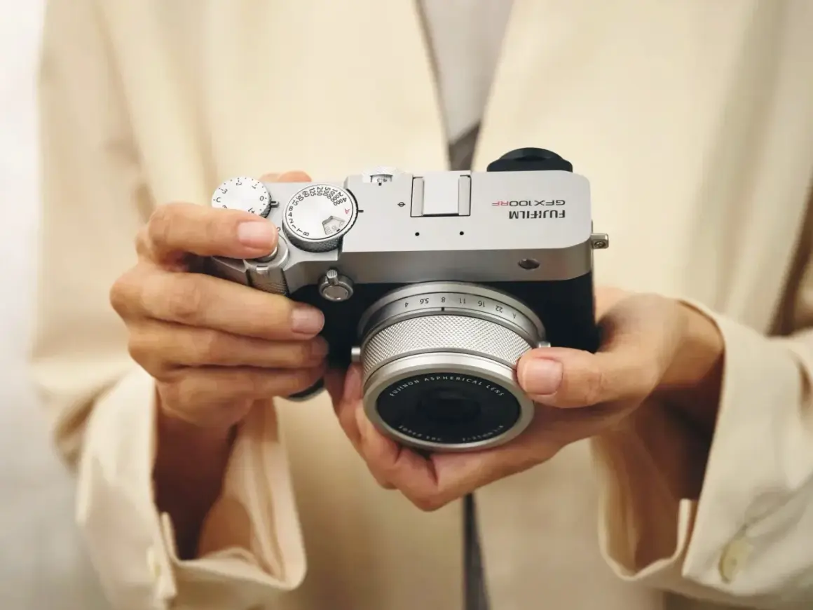 Händerna håller en silverfärgad Fujifilm GFX 100s kamera med ett objektiv.